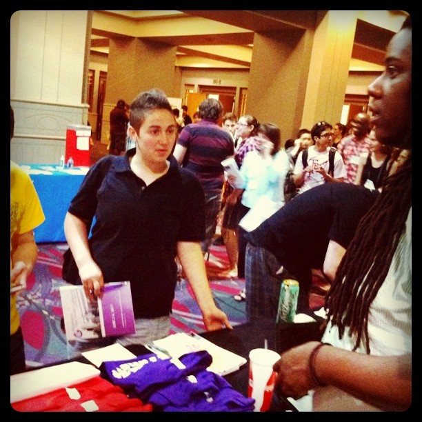 Asher Kolieboi, a black man with long locked hair stands behind a table speaking to a man with short brown hair wewaring a black polo shirt. Legalize Trans* shirts in red and purple are visible on the table.